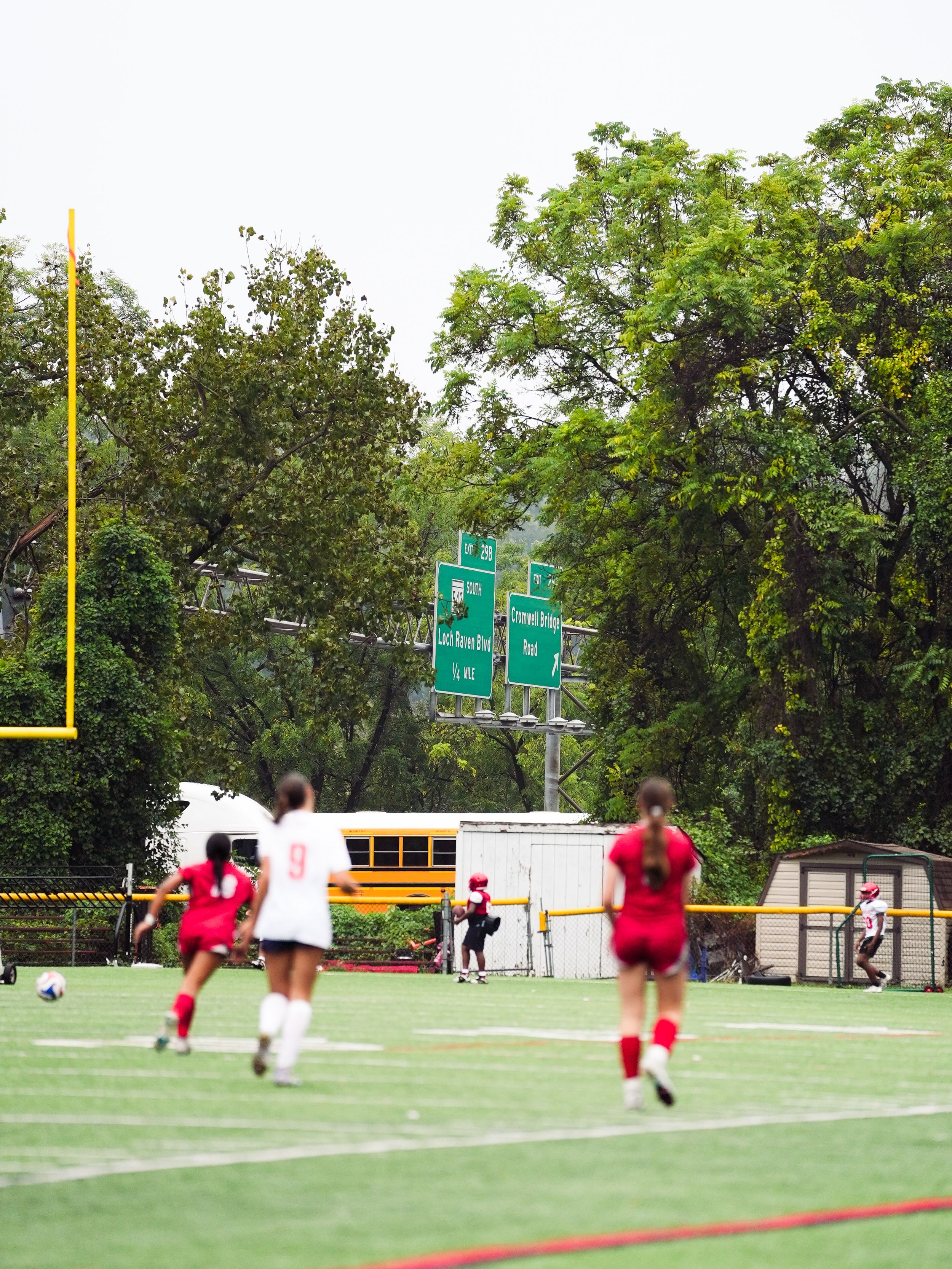 A sequence of football players practicing field goal kicks on a green practice field with goal posts and trees in background.