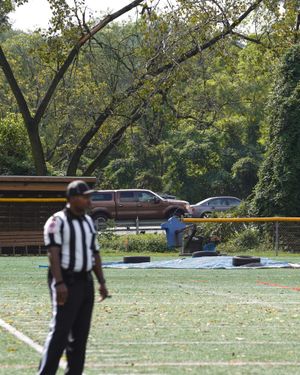 Referee stands on football field with trees and school bus visible in background.