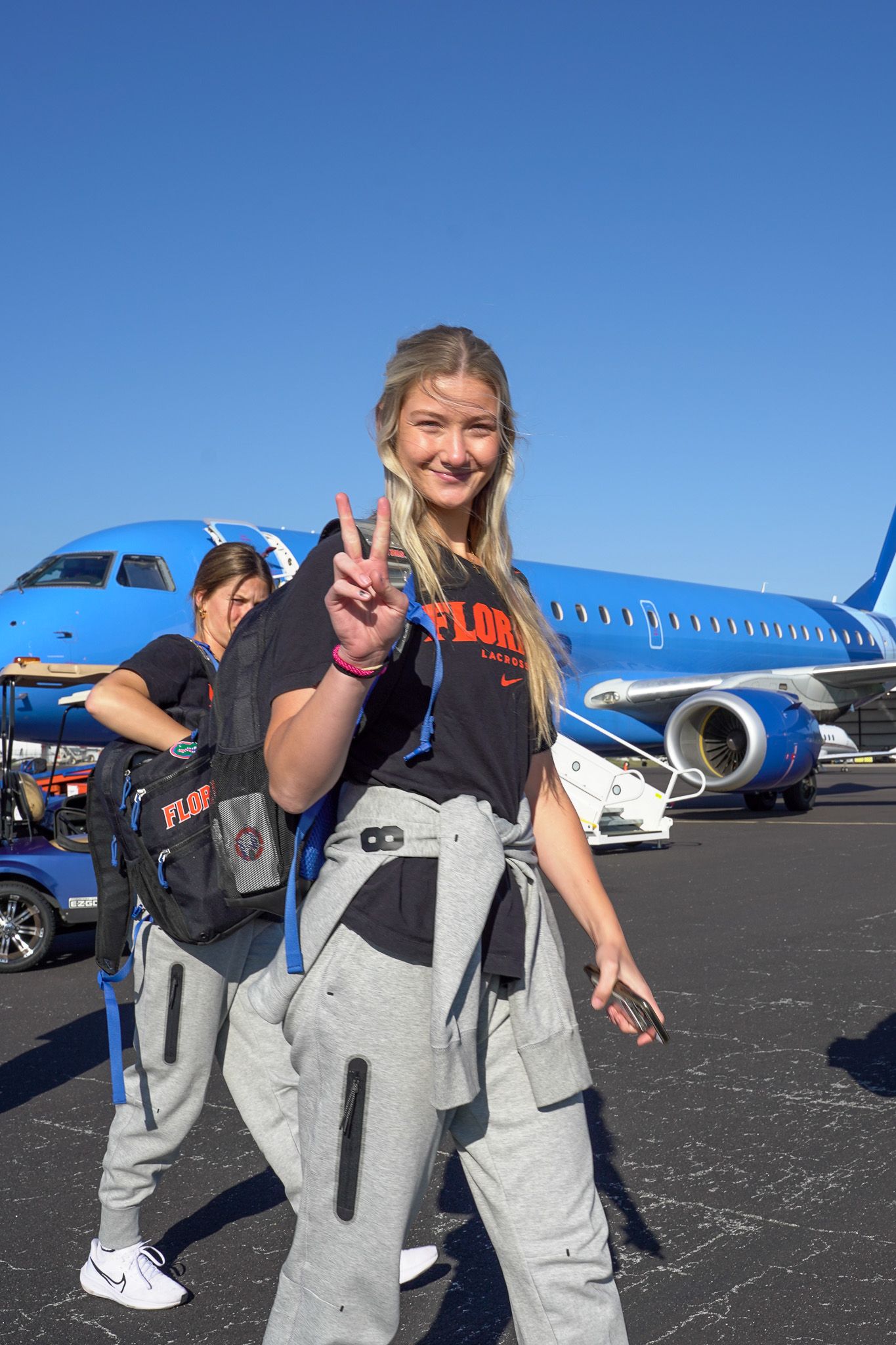 Student athlete poses with peace sign in front of blue aircraft on sunny day.