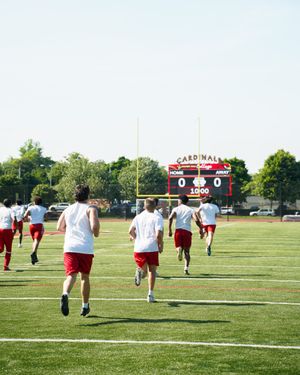Football players run towards scoreboard during practice on sunny field.