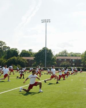 Football team performs stretching exercises on practice field under bright stadium lights.