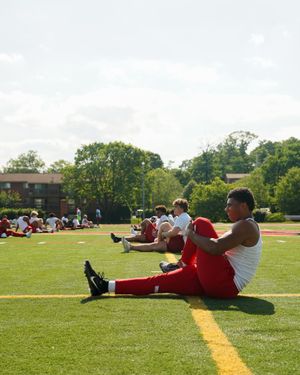 Athletes perform seated stretching exercises on football field sideline.