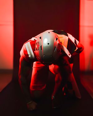 Football player in dramatic red lighting crouches in athletic stance wearing protective gear.