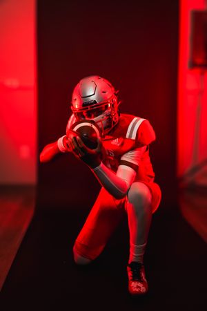 Football player poses in dramatic red lighting wearing helmet and uniform.
