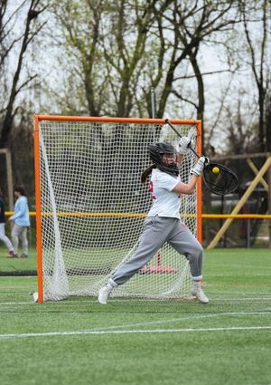 A lacrosse goalie in white and gray uniform positions defensively in front of the net on a grass field.