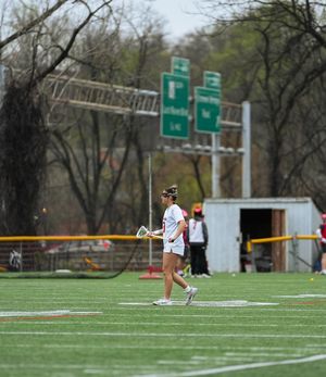 Lacrosse player in white uniform runs with stick on artificial turf field with green highway signs in background.