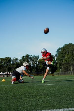 Football player kicks ball while another holds during field goal attempt.