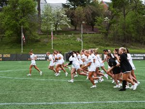 Two opposing lacrosse teams run across a field during a game on a bright spring day with trees in the background.