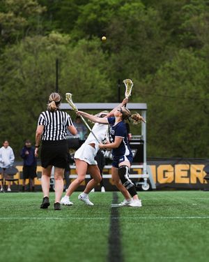 Lacrosse players competing for control of the ball during a match with a referee watching on an outdoor field.