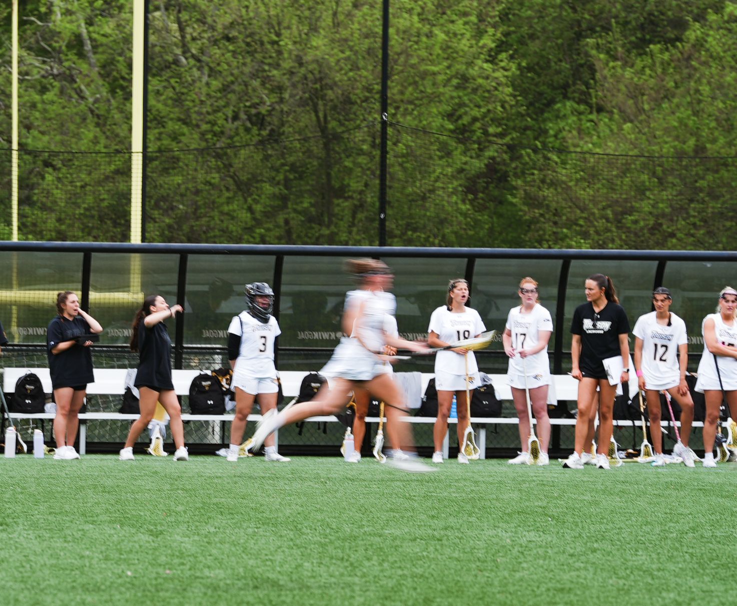 High school or college sports team watches from sideline as player runs past on artificial turf field during game.