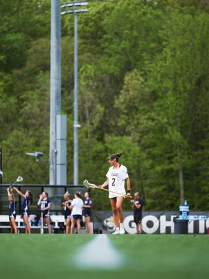 A lacrosse player in a white uniform leaps while holding their stick during an outdoor game with trees in background.