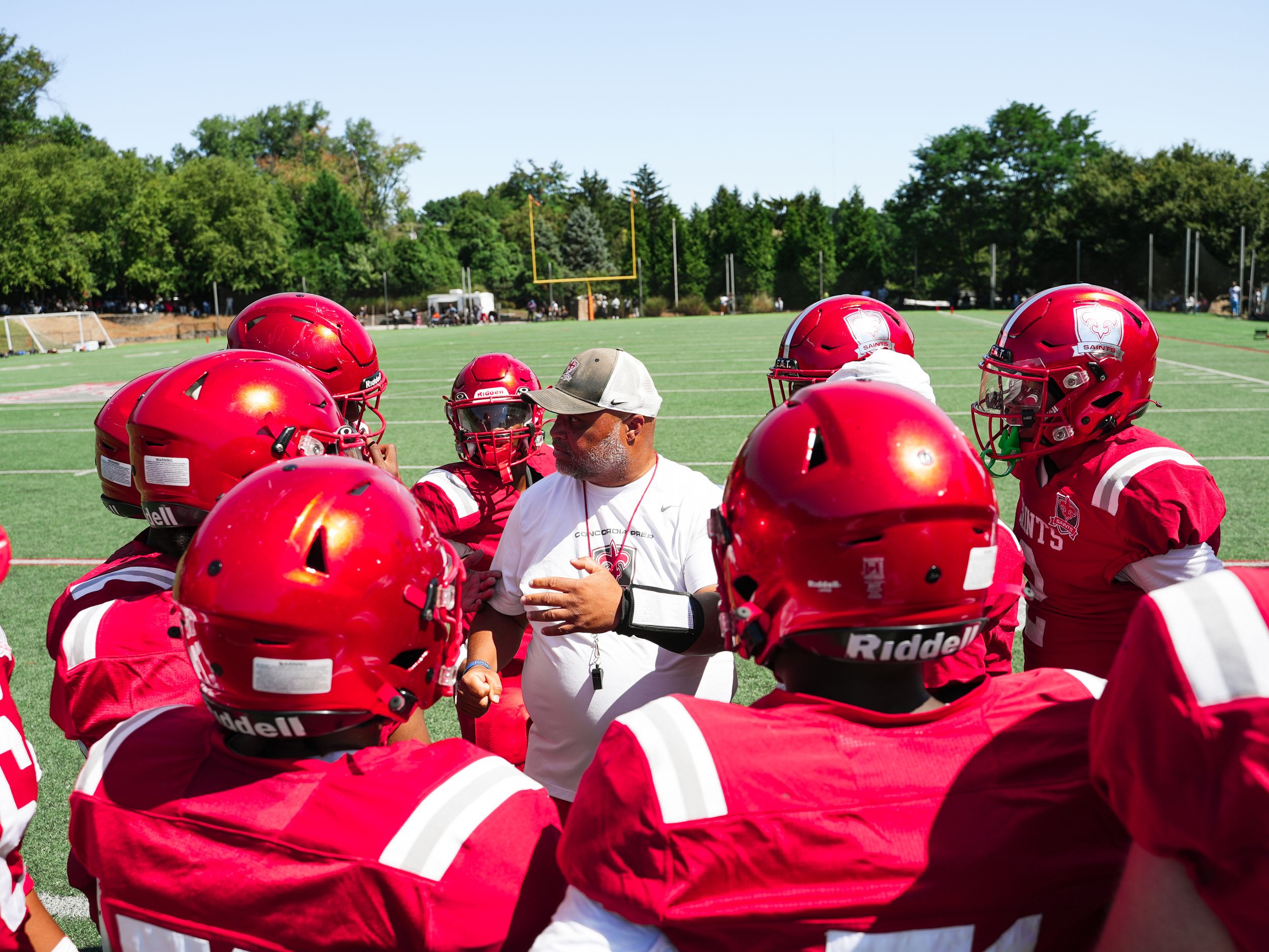 Football team huddles together wearing red jerseys during practice on a sunny day on the field.
