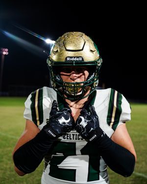 Football player in white Celtic jersey and gold helmet posing under stadium lights at night.