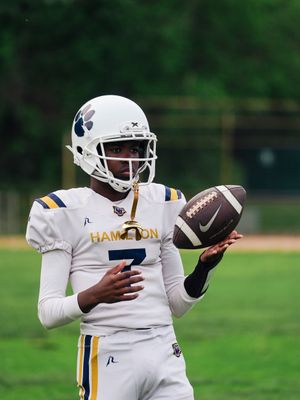 Football player in white Hampton uniform number 7 holds ball during game with green background.