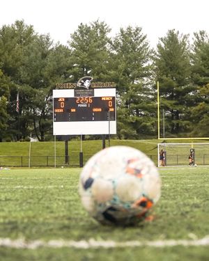 Soccer ball in foreground with scoreboard and trees visible in background.