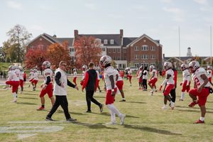 Football players practicing drills on field with brick buildings in background.