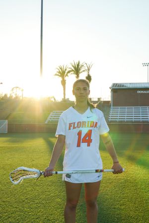University of Florida athlete wearing number 14 stands with lacrosse stick at sunset on athletic field.