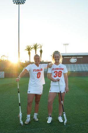 Two Florida Gators lacrosse players in white uniforms pose together on field at sunset.