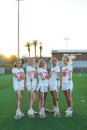 Five Florida Gators lacrosse players in white uniforms pose with sticks during sunset photoshoot.