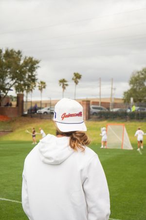 Spectator in white hoodie and baseball cap watching sports game from sideline.