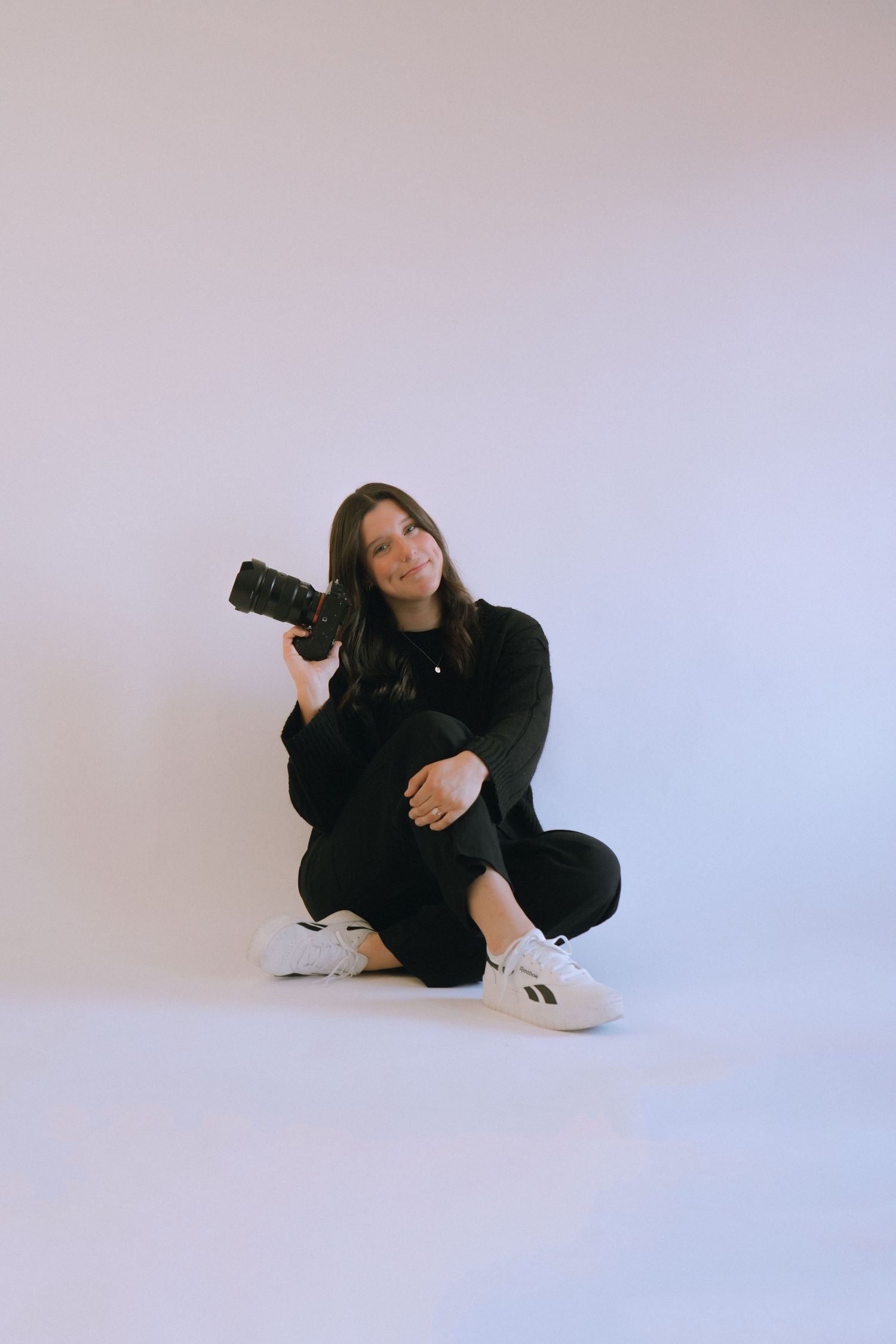 Photographer in all black outfit sits with camera against white studio background.