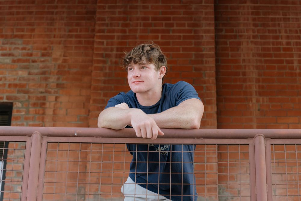 Casual portrait of someone leaning on a wooden railing against brick wall background.