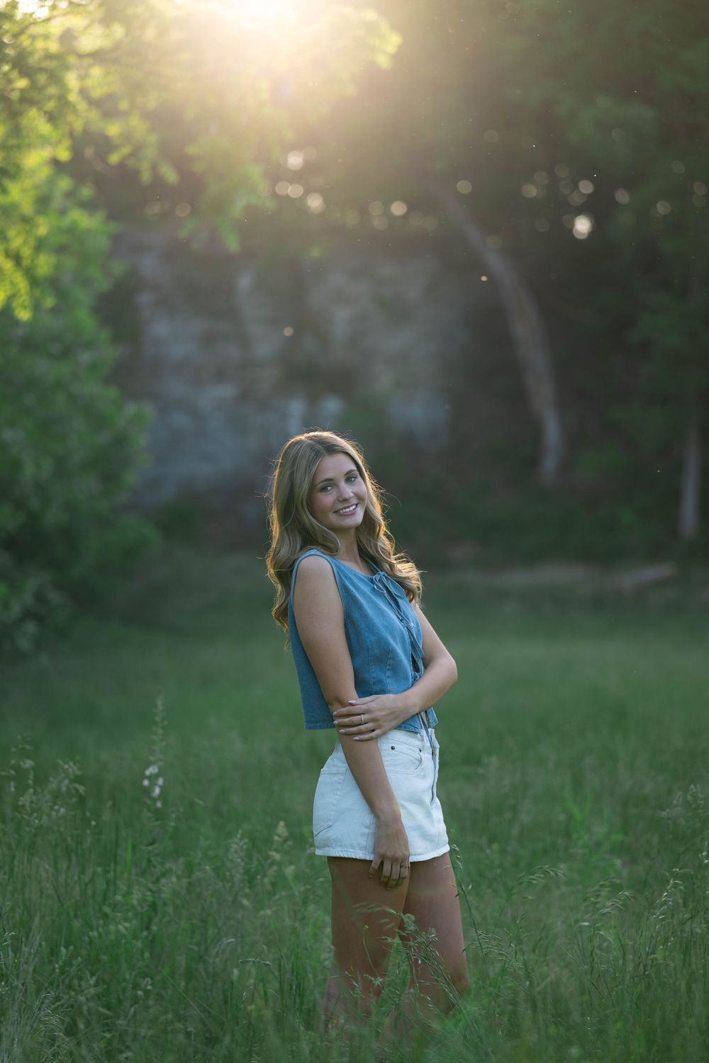 Young woman in denim top and white shorts smiling in sunlit green field with golden hour backlight.