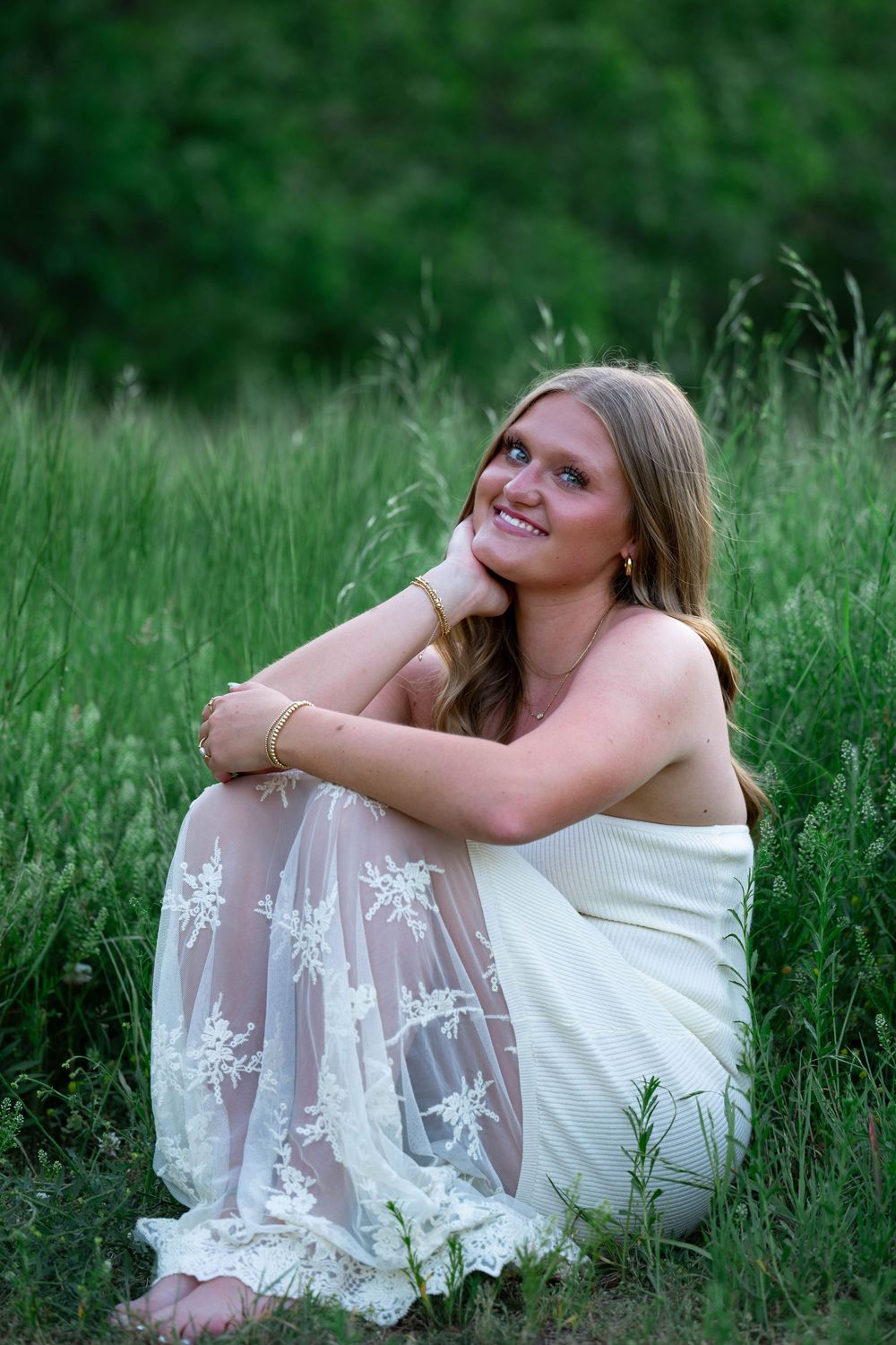 Young woman in white lace dress sitting in lush green grass smiling with hand on chin.