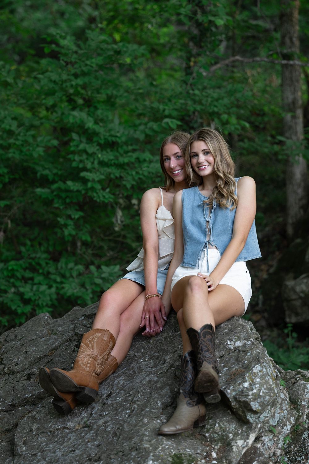 Two young women in cowboy boots and summer outfits smile while sitting together on a large rock in a forest.