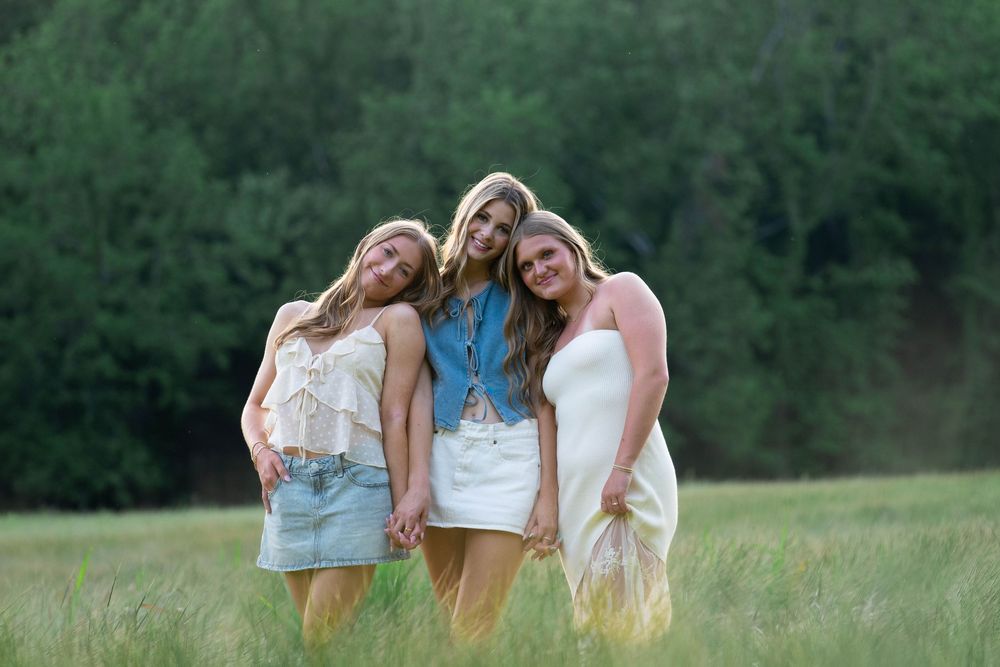 Three young women in summer outfits smile and pose together in a green meadow with trees in the background.