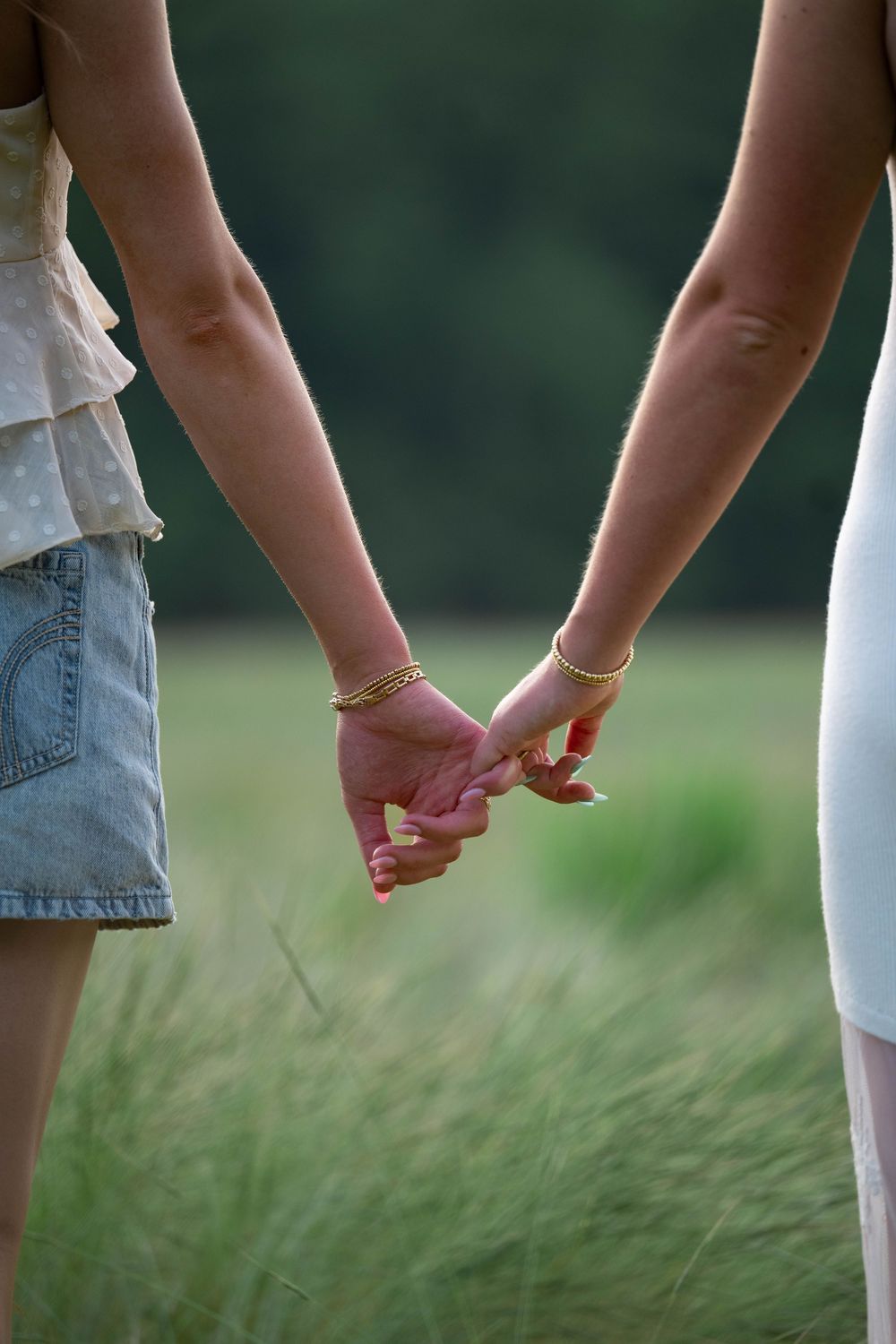 Two women holding hands with gold bracelets in a green field, symbolizing friendship or love.