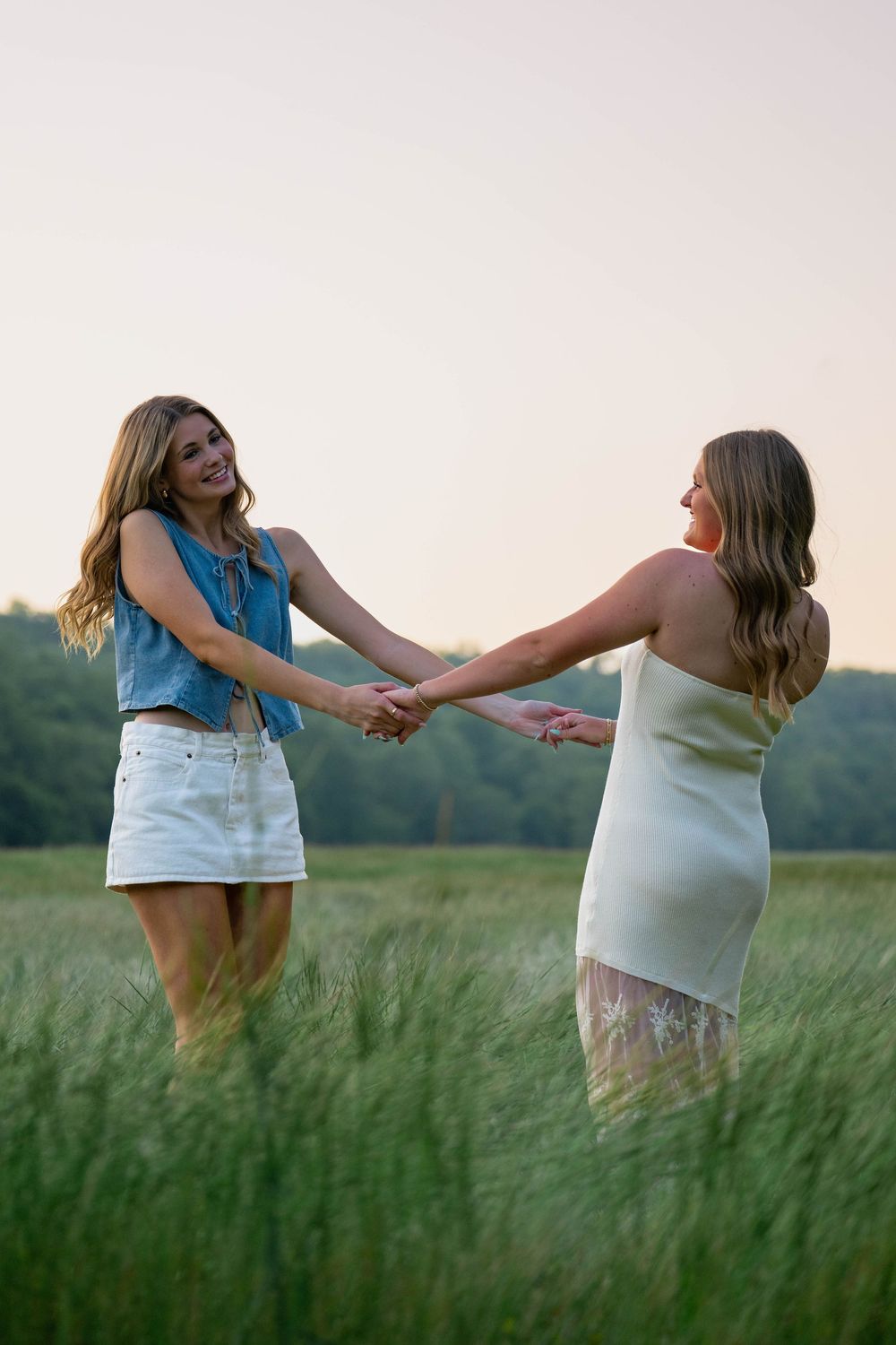 Two young women in white and denim outfits hold hands and spin joyfully in a lush green field at sunset.