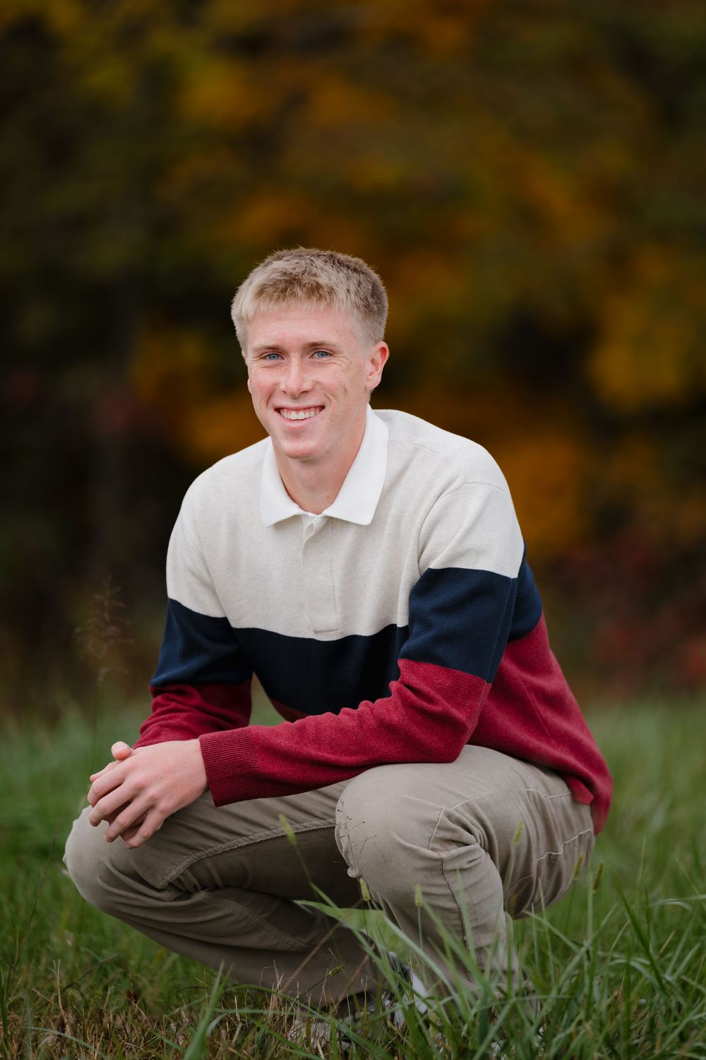 Young man in colorful sweater crouching in a grassy field with autumn foliage in the background.