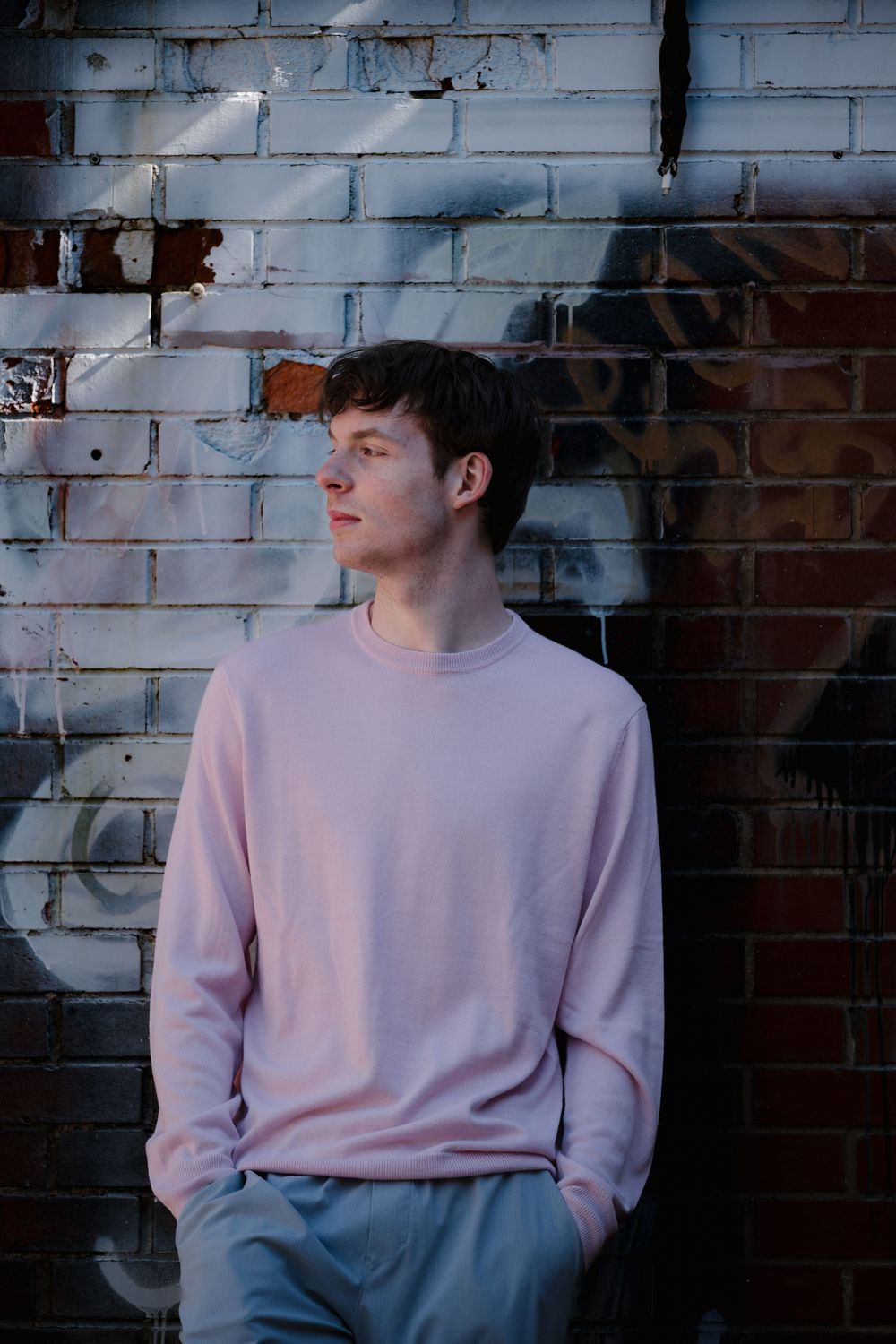 Young man in pink sweatshirt and gray pants leaning against a graffiti brick wall outdoors.