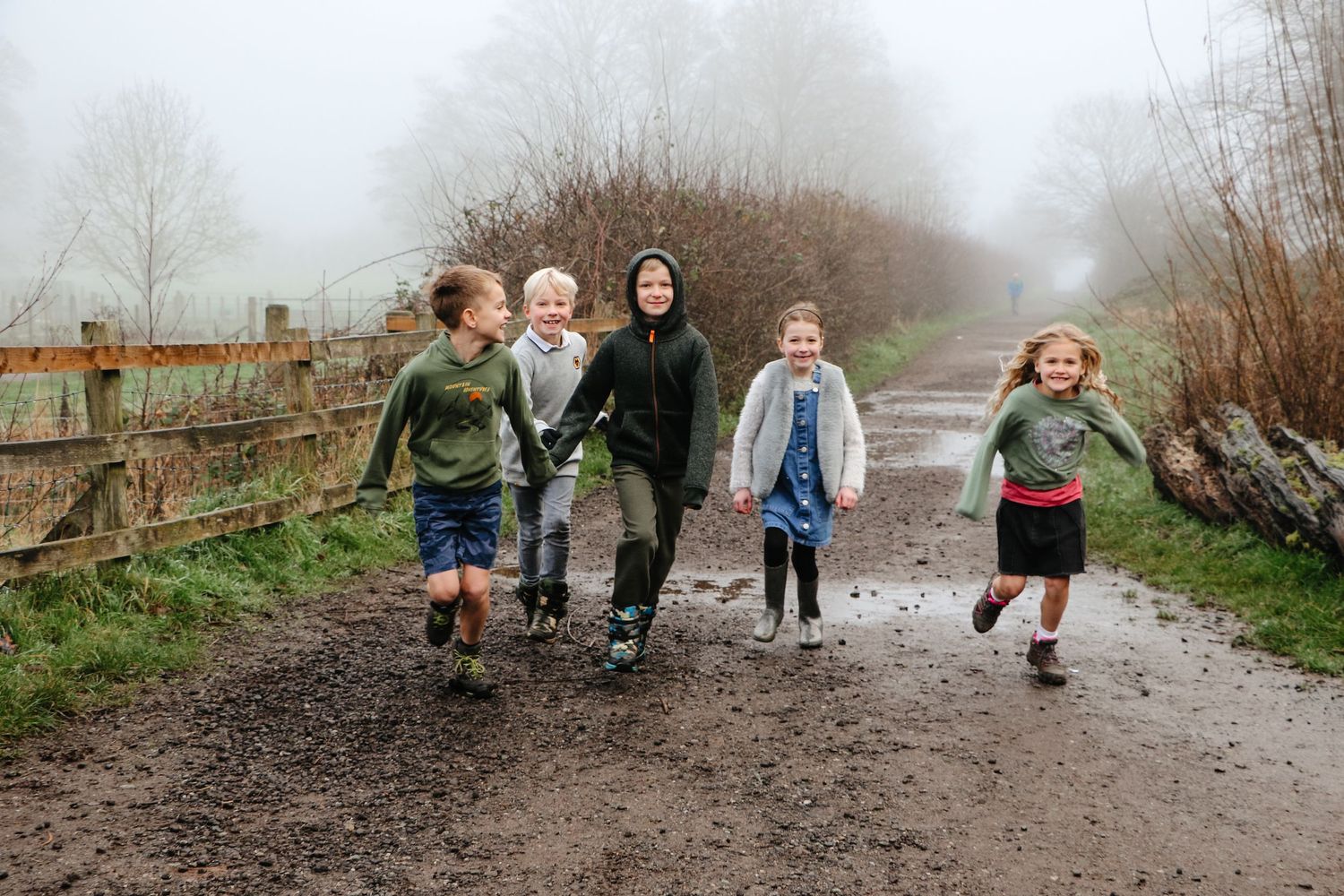 Children run joyfully down a muddy path on a misty day in the countryside.