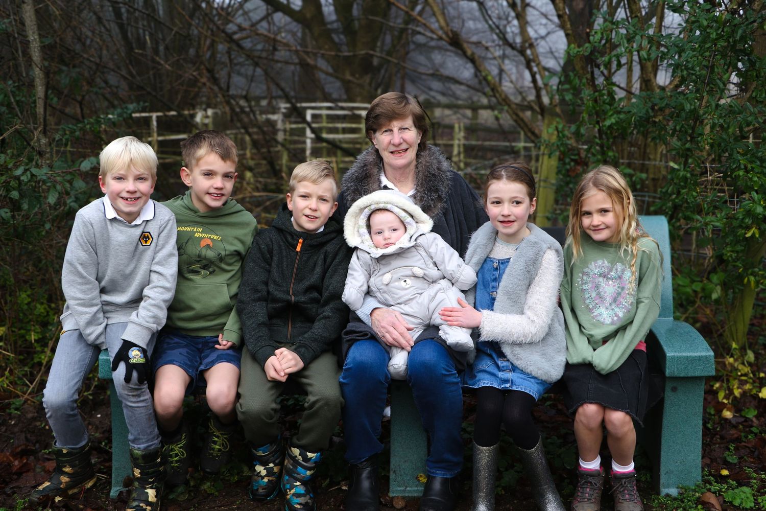 A group of children sitting together on a green bench in a garden setting, including a small baby in winter clothes.