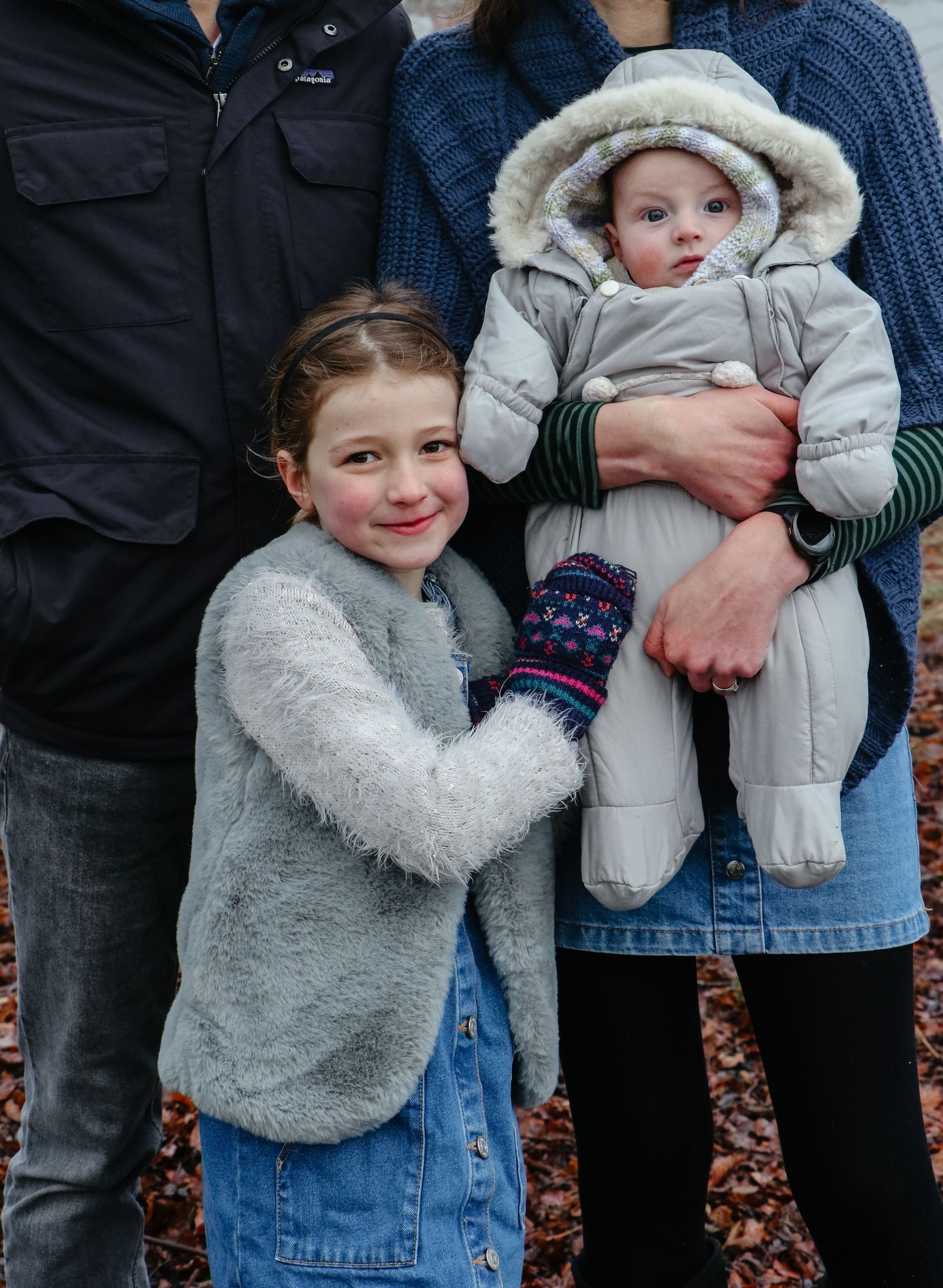 Two people in winter clothing standing together outdoors with a baby wearing a hooded snowsuit.