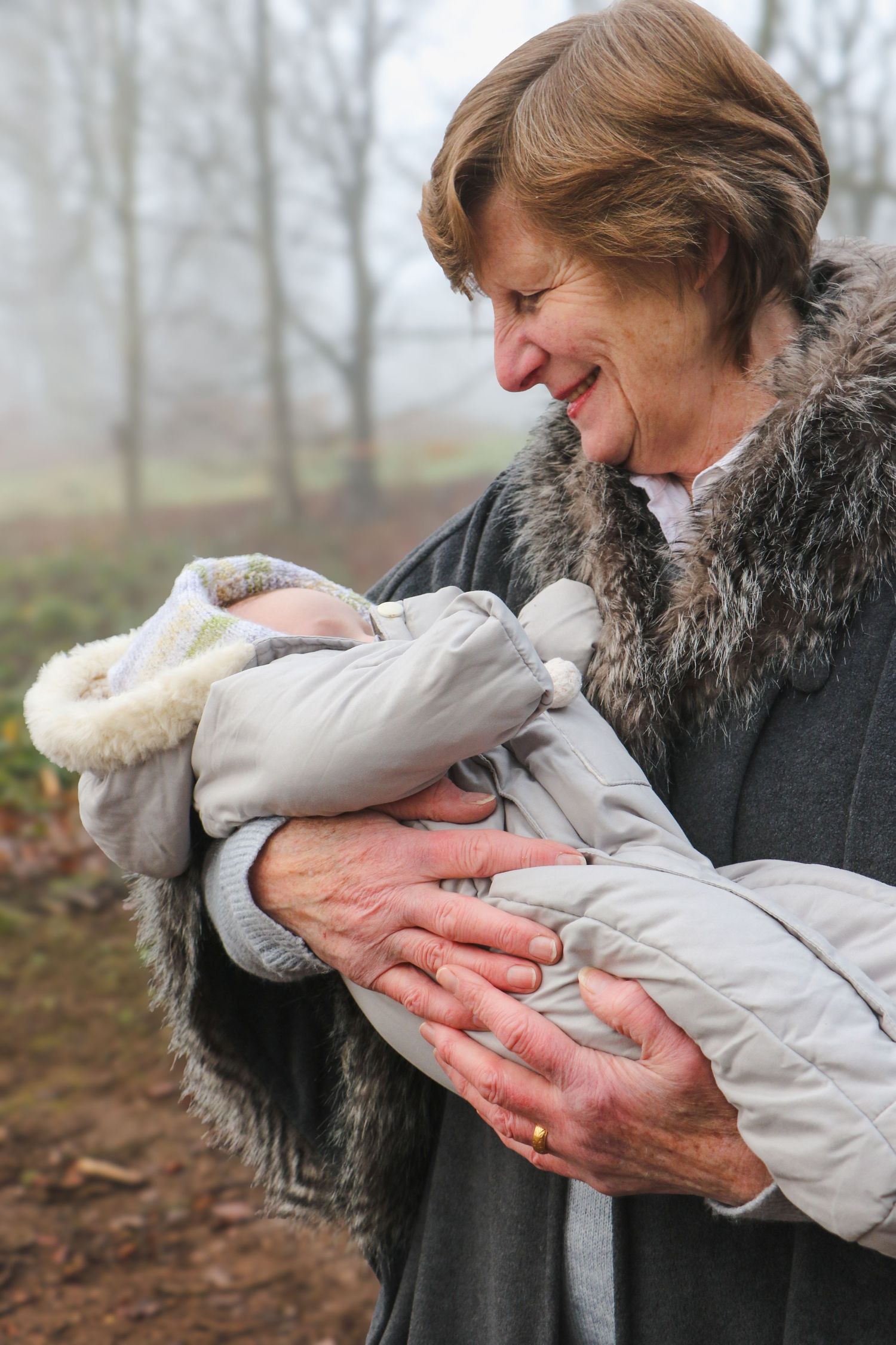 A person in a fur-trimmed coat smiles warmly while holding a bundled baby on a misty autumn day.