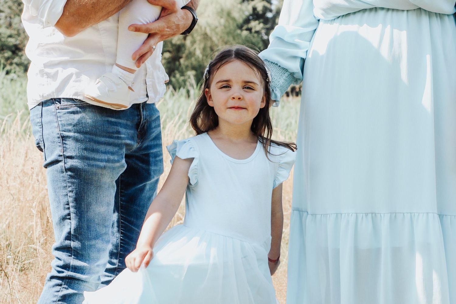 A child in a white dress standing outdoors among family members on a sunny day.