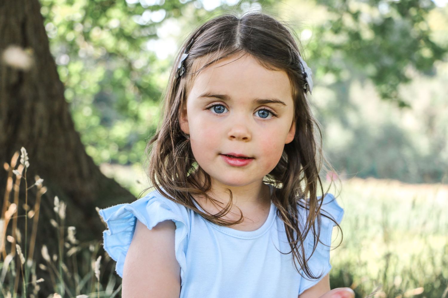 A young child in a light blue dress poses outdoors in natural sunlight.