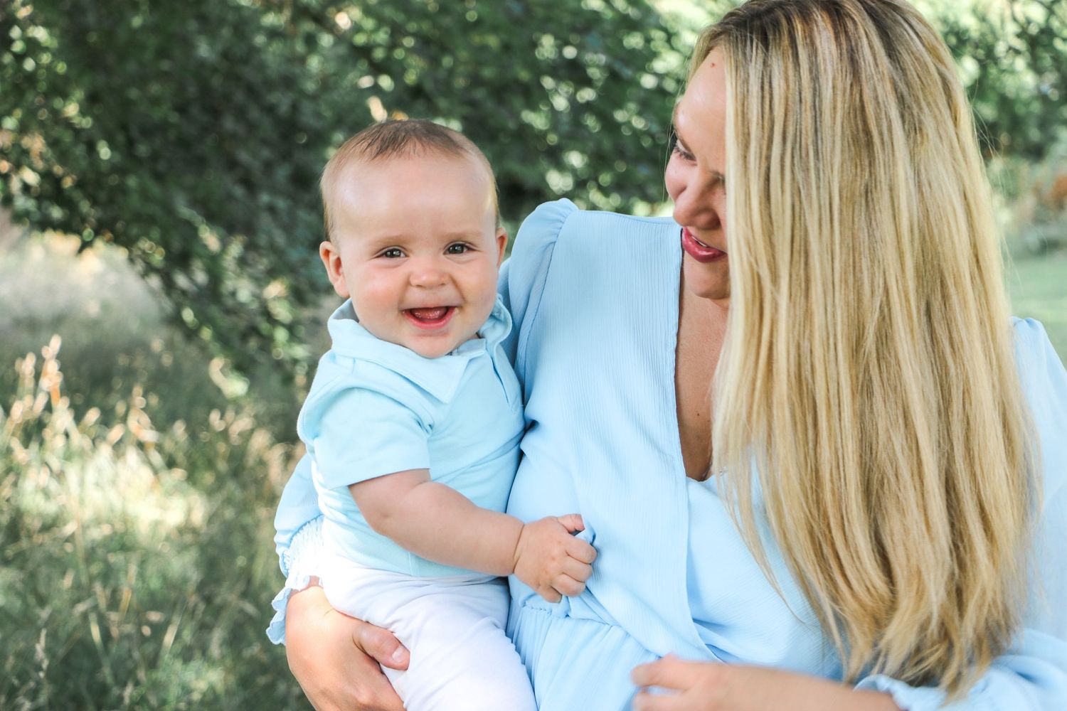 A baby in light blue clothing shares a happy moment outdoors.