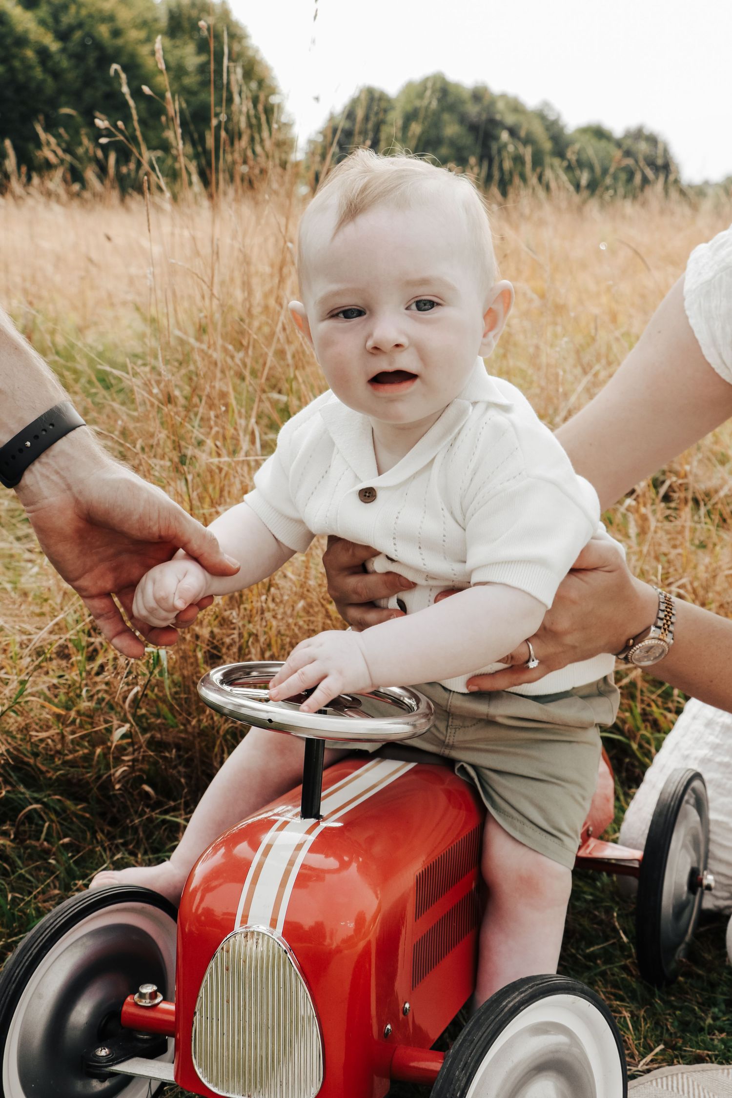 A baby sits on a vintage red toy car in a field of tall grass.
