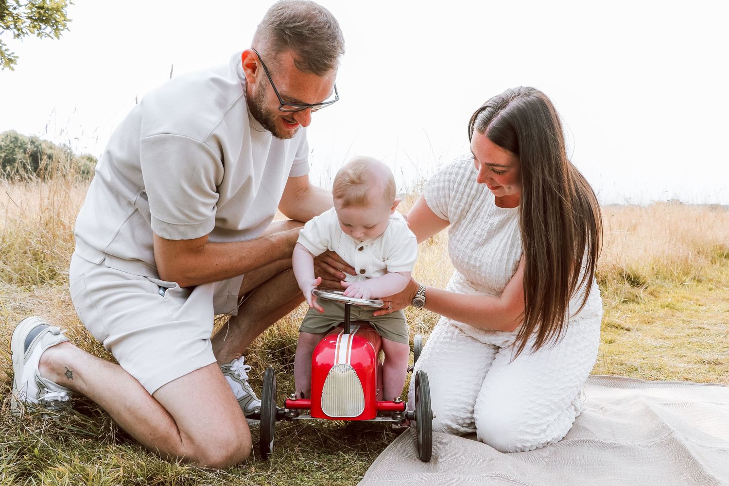 Parents help their baby explore a red toy car during an outdoor photoshoot.
