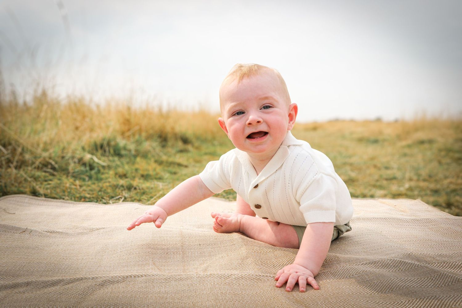 Smiling baby in white outfit crawls on beige blanket outdoors with golden field in background.