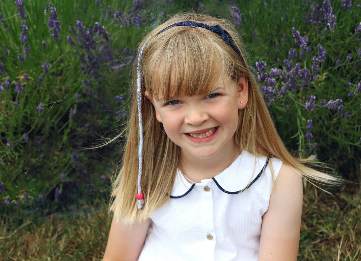 A child in a white dress with a peter pan collar stands in a garden with purple wildflowers blooming in the background.