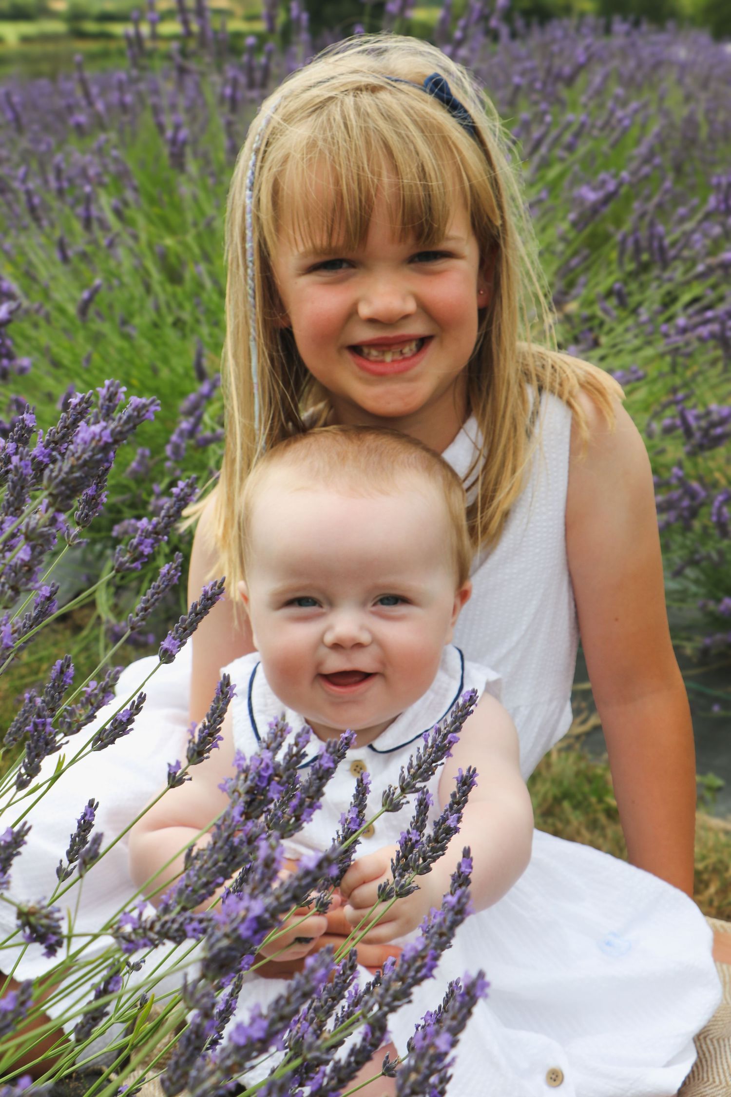 Two children in white dresses pose together among blooming purple lavender flowers in a garden.