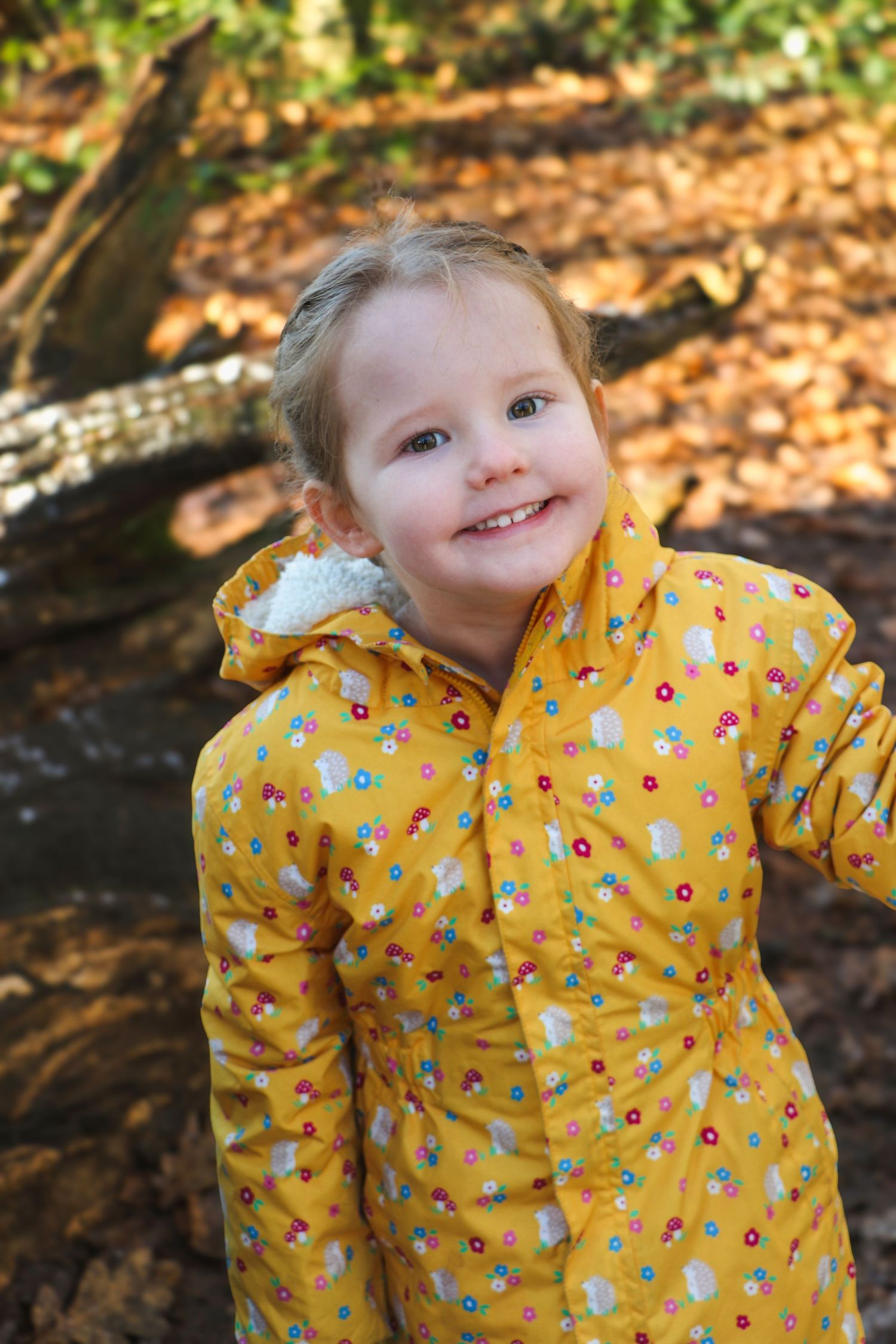 Child wearing a bright yellow floral raincoat plays outdoors among fallen autumn leaves.