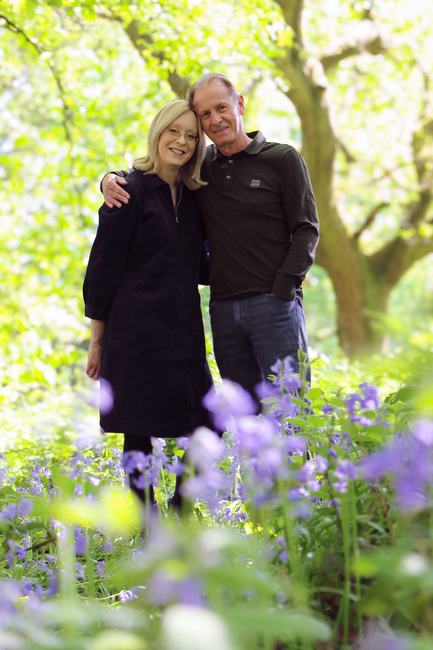 A couple stands together smiling among purple wildflowers in a sun-dappled spring forest setting.