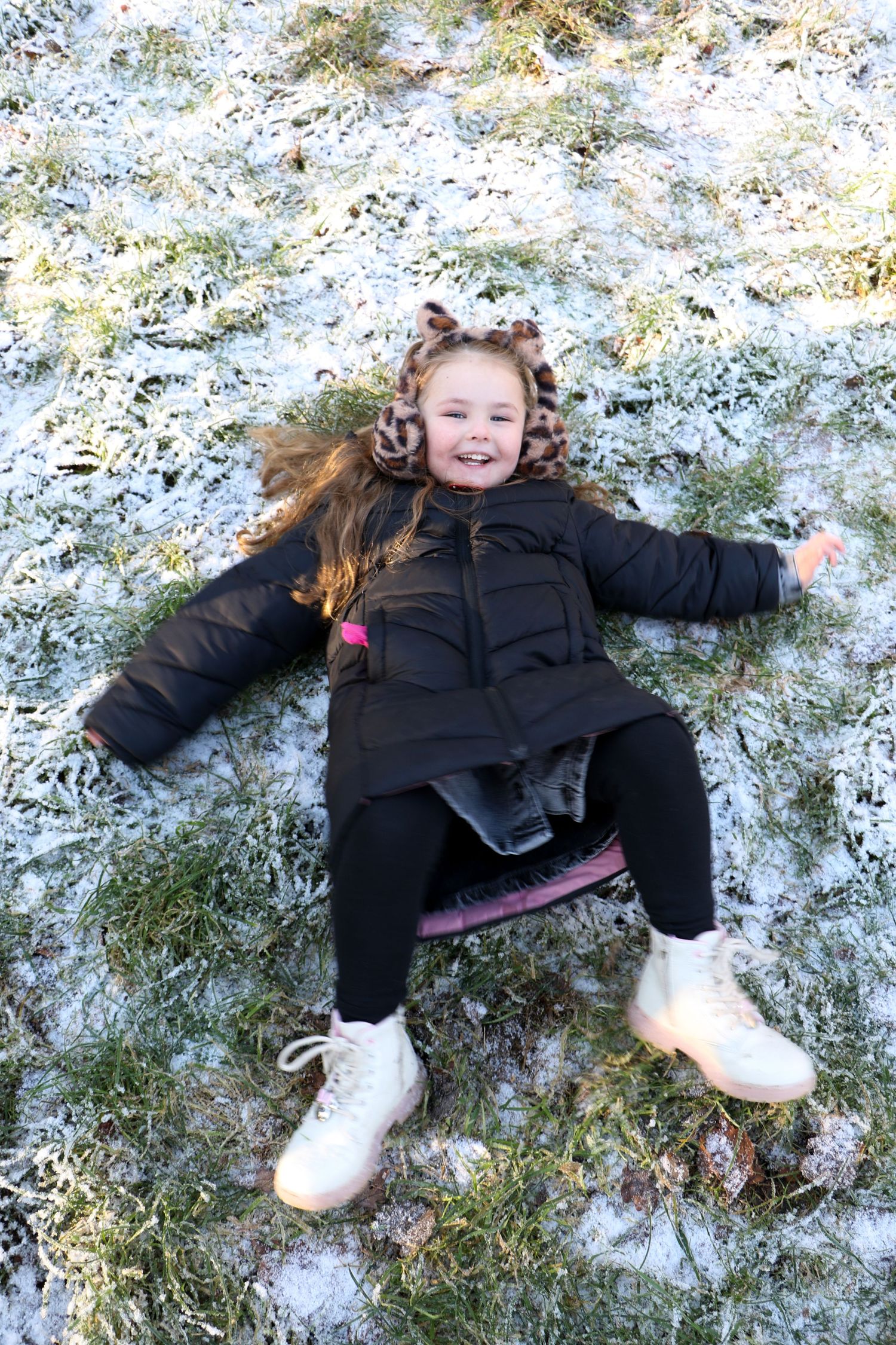 A child in a black winter coat lies making snow angels in fresh powder snow with joyful expression.