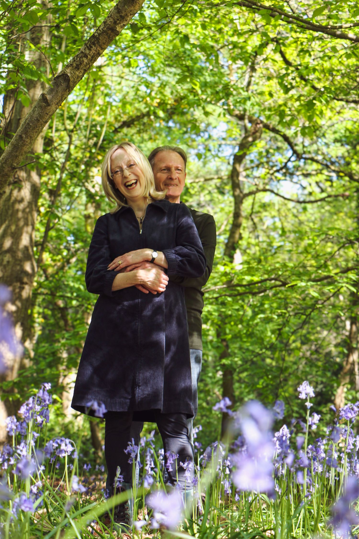A couple embraces while standing in a woodland garden with blooming purple bluebells in the foreground.